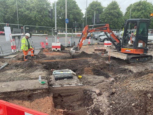 A photo of a small digger digging a hole, the area is uneven and has piles of soil. A workman in high vis is stood with a leg in a hole.