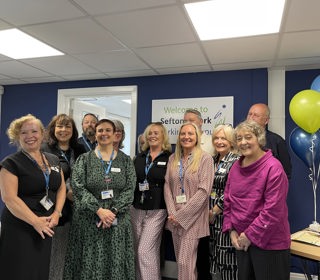 Group of people from the Sefton@Work team and Cllr Paulette Lappin in an office setting, smiling. Celebratory balloons are on the desk. 
