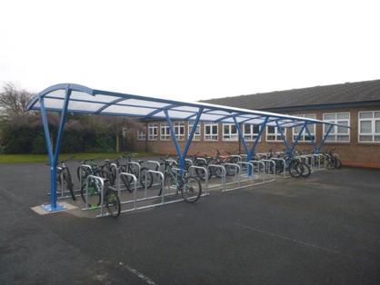 A slanted angle of a bike shelter next to a school. It has blue supports and a plastic roof, with room for around 40 bikes.
