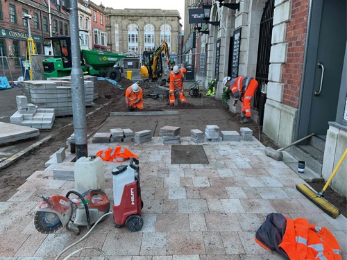 A progress picture of King Street, three workmen in high vis are preparing the ground for pavement tiles, half the street has new paving tiles. There are piles of more paving tiles. - January 2025