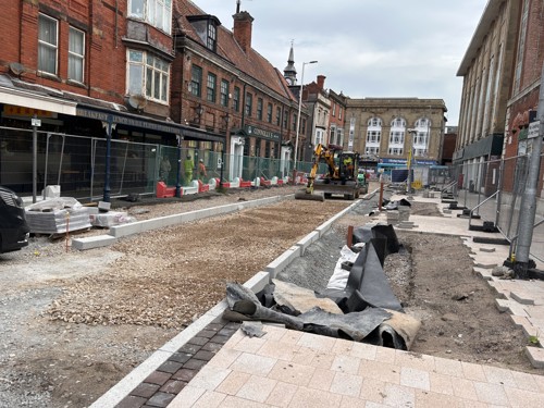 Gravel being flattened by a roller on King Street, with pavement kerbs put in and most of the paving slabs on the right market side complete. There are big bits left unpaved for a future rain garden.